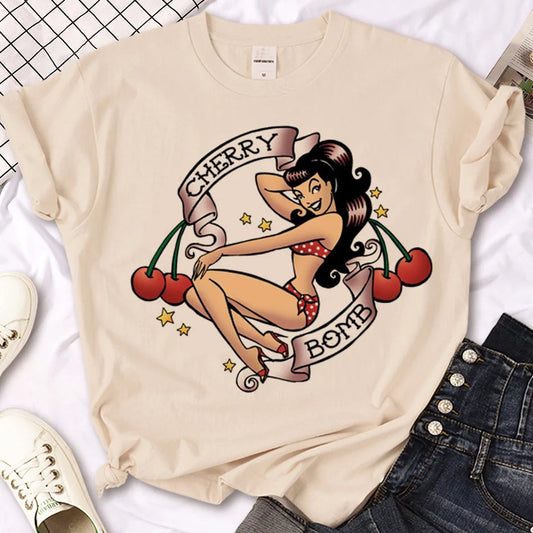 Beige t-shirt with a graphic design of a woman and cherries on a white surface with shoes and jeans.