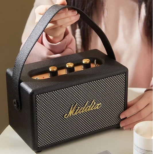 Black portable speaker with gold accents on a table, person holding it, candle in the background

