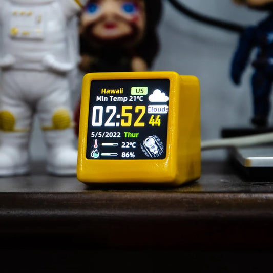 Digital clock with weather display on a wooden surface, with figurines in the background.